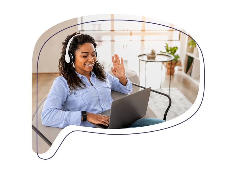 Eine Frau mit Headset sitzt mit einem Laptop auf dem Schoß und winkt lächelnd in eine Videokamera.