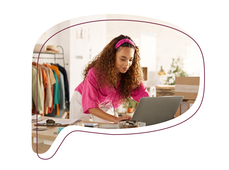 Eine Frau arbeitet konzentriert an einem Laptop in einem hellen Atelier, im Hintergrund hängen bunte Kleidungsstücke auf einer Stange.