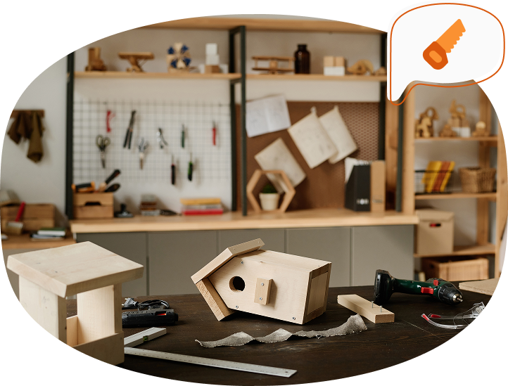 Blick in eine Schreinerwerkstatt: Auf einem Arbeitstisch liegen ein selbst gebautes Vogelhaus, Werkzeug und Holzteile, im Hintergrund Regale mit Materialien und Werkzeugen.
