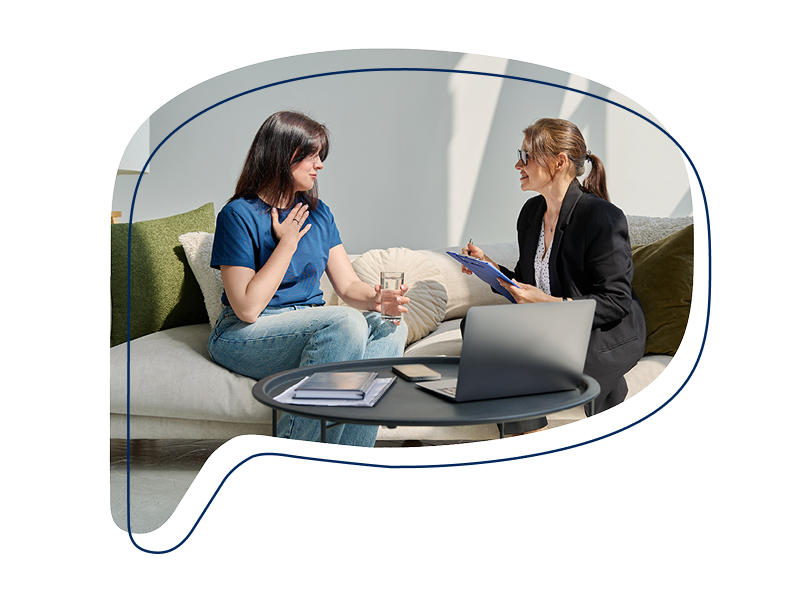 Zwei Frauen sitzen auf einem Sofa im Gespräch – eine hält ein Glas Wasser und spricht, die andere macht sich Notizen auf einem Klemmbrett.